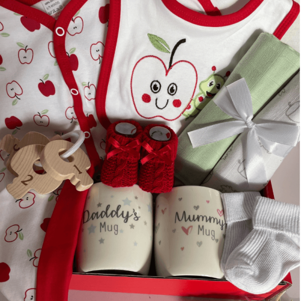 Apple Baby Gift Set, a white gift box thats filled with a baby clothing set with a bright red apple print on, matching red knit booties, 2 muslin squares and some mugs for mum and dad